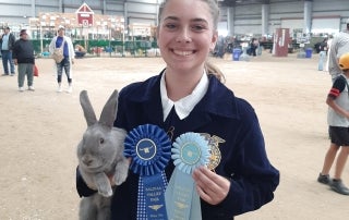 A young women, in a blue FFA jacket, holding a grey rabbit and two blue ribbons. She smiles proudly, and is standing in the middle of the barn, with goat pens behind her in the distance.
