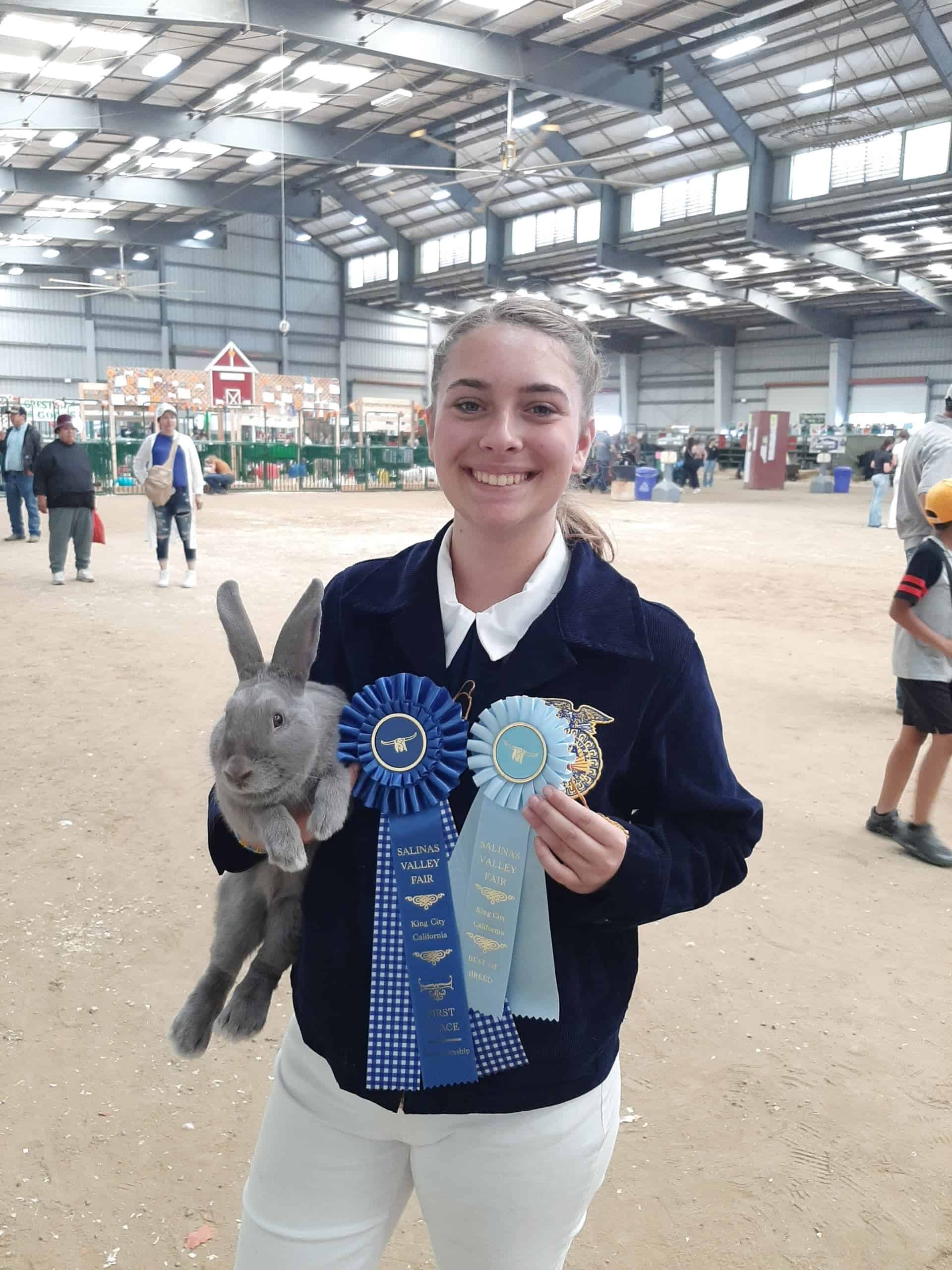 A young women, in a blue FFA jacket, holding a grey rabbit and two blue ribbons. She smiles proudly, and is standing in the middle of the barn, with goat pens behind her in the distance.
