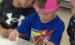 Shattuck Kindergarten student, Westin Burns, practicing giving shots at the vet science station with the help of Shattuck FFA Senior Avery Quisenberry while wearing his ear notched headband from an ear notching station.