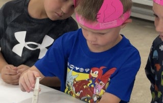 Shattuck Kindergarten student, Westin Burns, practicing giving shots at the vet science station with the help of Shattuck FFA Senior Avery Quisenberry while wearing his ear notched headband from an ear notching station.