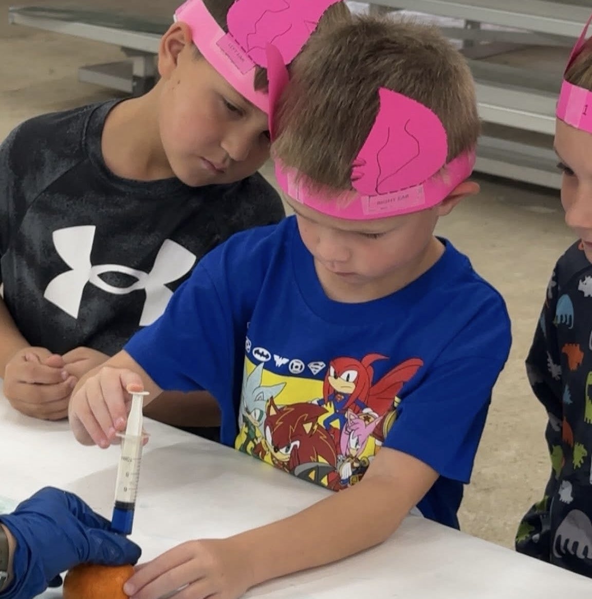 Shattuck Kindergarten student, Westin Burns, practicing giving shots at the vet science station with the help of Shattuck FFA Senior Avery Quisenberry while wearing his ear notched headband from an ear notching station.