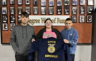 Three Barnesville FFA Freshman–Caleb Bush, Chloe Gordon, and Aiden Starr–stand in front of the Degree Hall of Fame holding their new blue FFA jacket embroidered with "Ohio" and "Barnesville."
