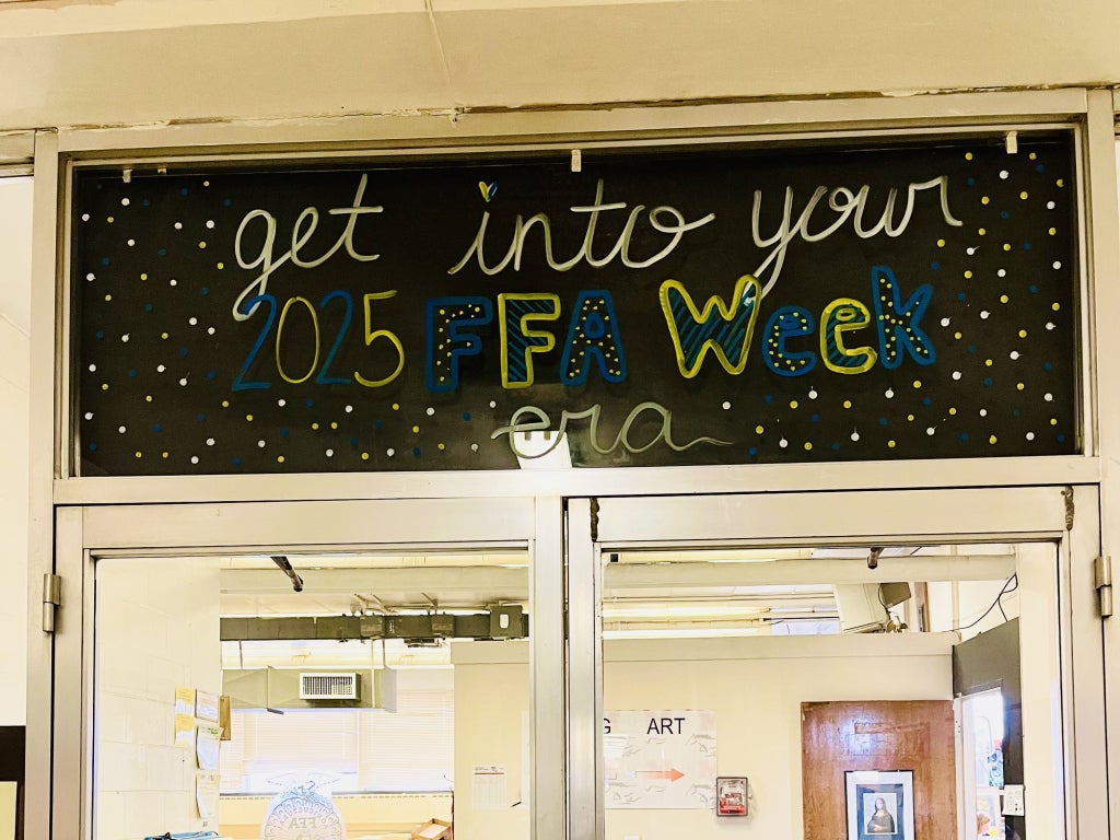 A decorated school hallway sign reads “Get into your 2025 FFA Week era” in blue and gold colors above classroom doors.