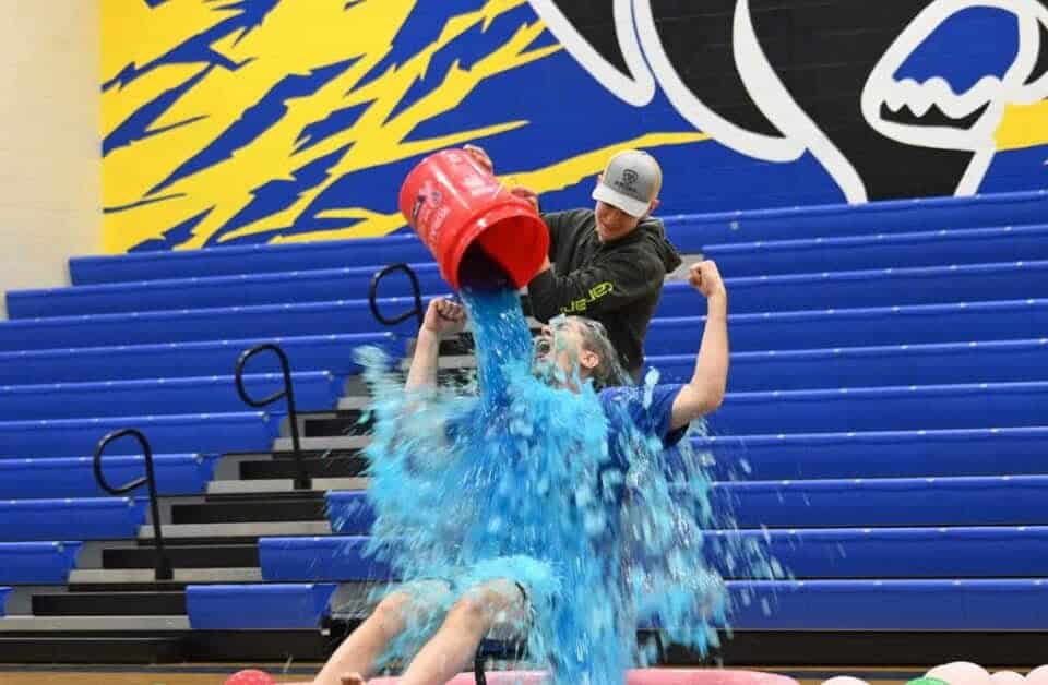 A student pours a bucket of blue slushie over a teacher during a school fundraiser in a gymnasium.
