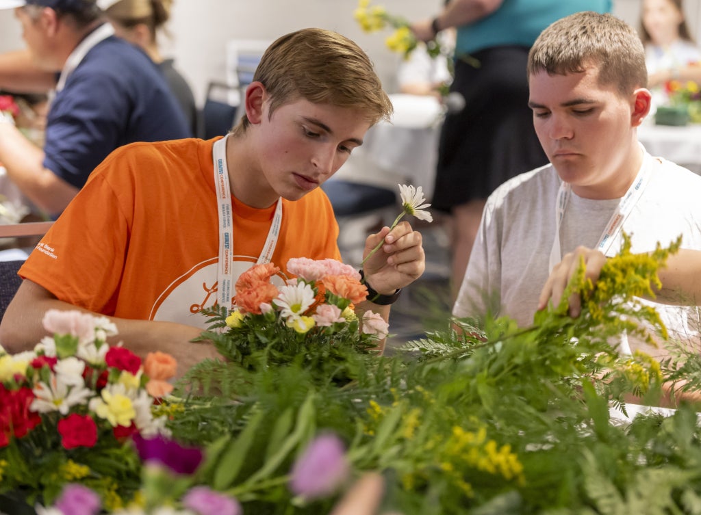 Two young people in casual attire engage in floral arrangement during an FFA educational workshop, surrounded by fresh flowers and greenery at an indoor conference setting in June 2026.