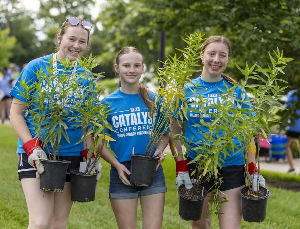 Three young members of the FFA wearing blue t-shirts participate in an outdoor tree-planting activity at the 2026 Catalyst Conference. Each student holds a small tree sapling in a black container as part of what appears to be an environmental service project in a park setting.