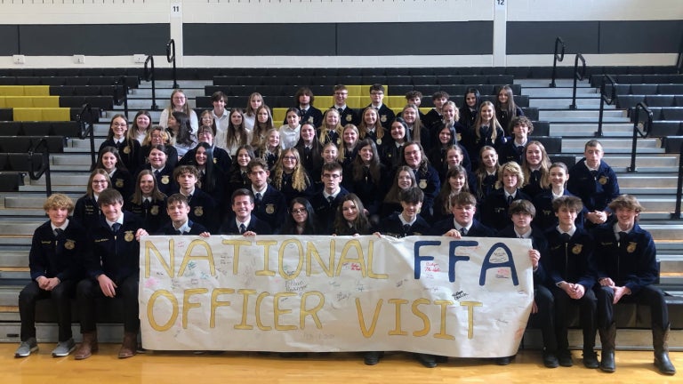 FFA members sit in official dress holding a sign that says "national FFA officer visit"