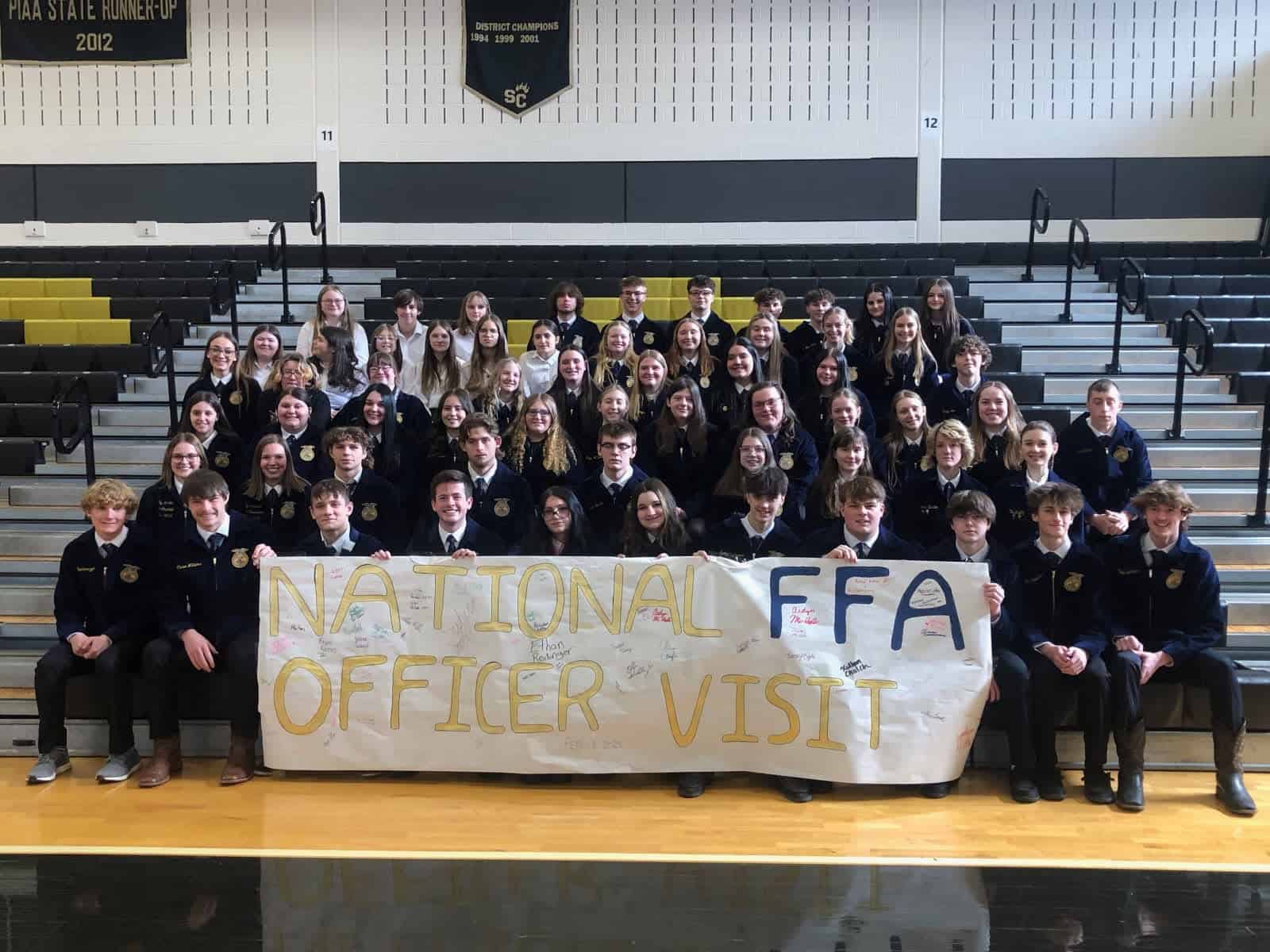 FFA members sit in official dress holding a sign that says "national FFA officer visit"