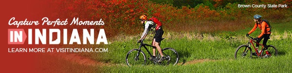 Advertisement for Visit Indiana reading “Capture Perfect Moments in Indiana — Learn more at VisitIndiana.com,” with a photo of two cyclists wearing helmets riding mountain bikes along a grassy trail at Brown County State Park, surrounded by trees with autumn red foliage.