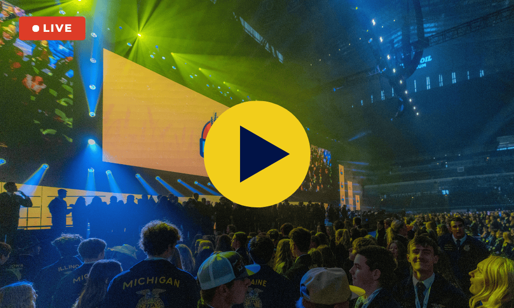 Live event photo from the 98th National FFA Convention & Expo showing a crowded arena filled with attendees, many wearing FFA jackets with state names. A large stage with yellow lighting and an FFA logo screen is in the background, lit by green and blue spotlights. A “LIVE” badge appears in the top left corner, and a yellow play button icon is overlaid in the center of the image, indicating a video.